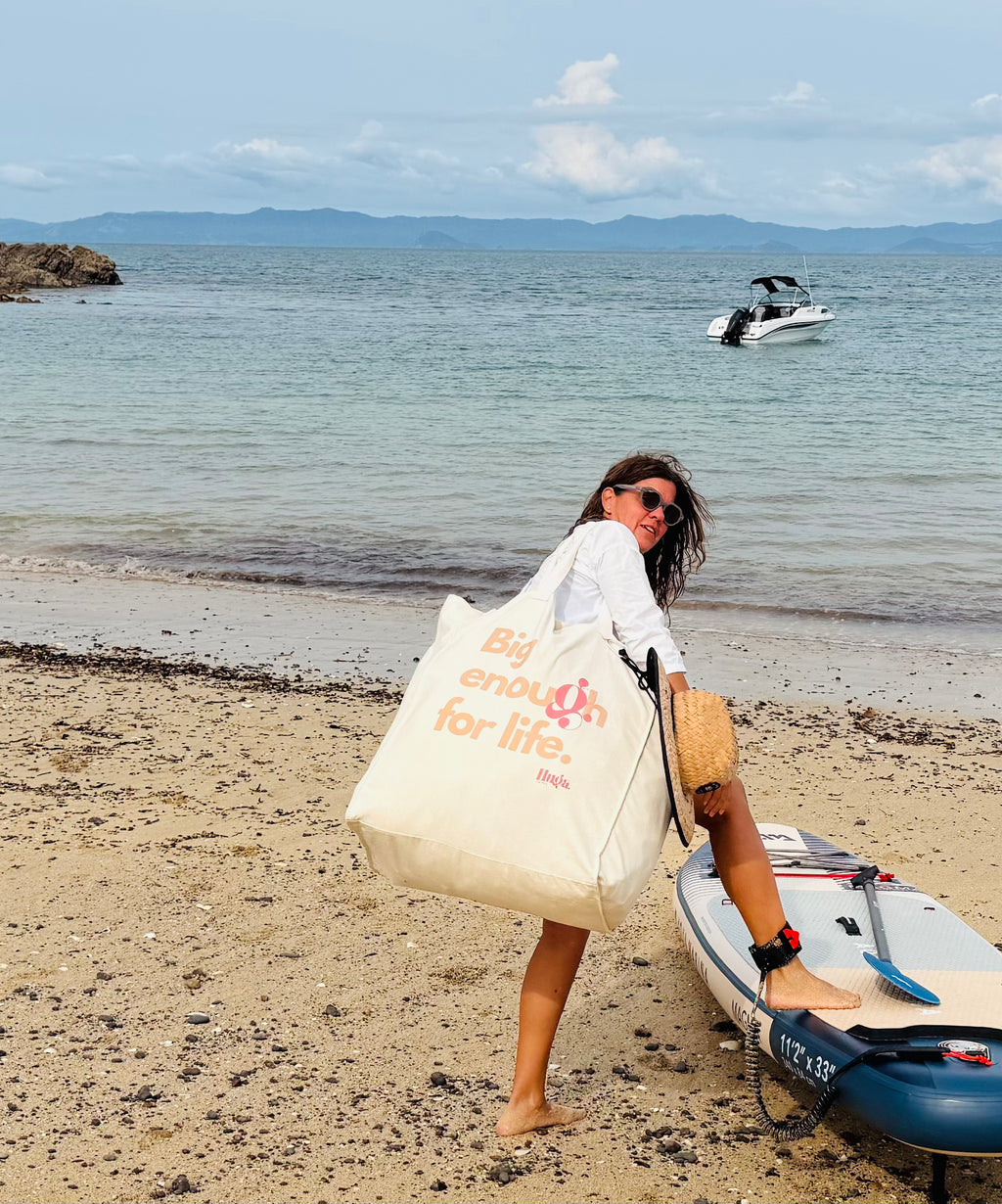 Woman on a beach with a paddleboard, holding a tote bag with text, and a boat in the background.
