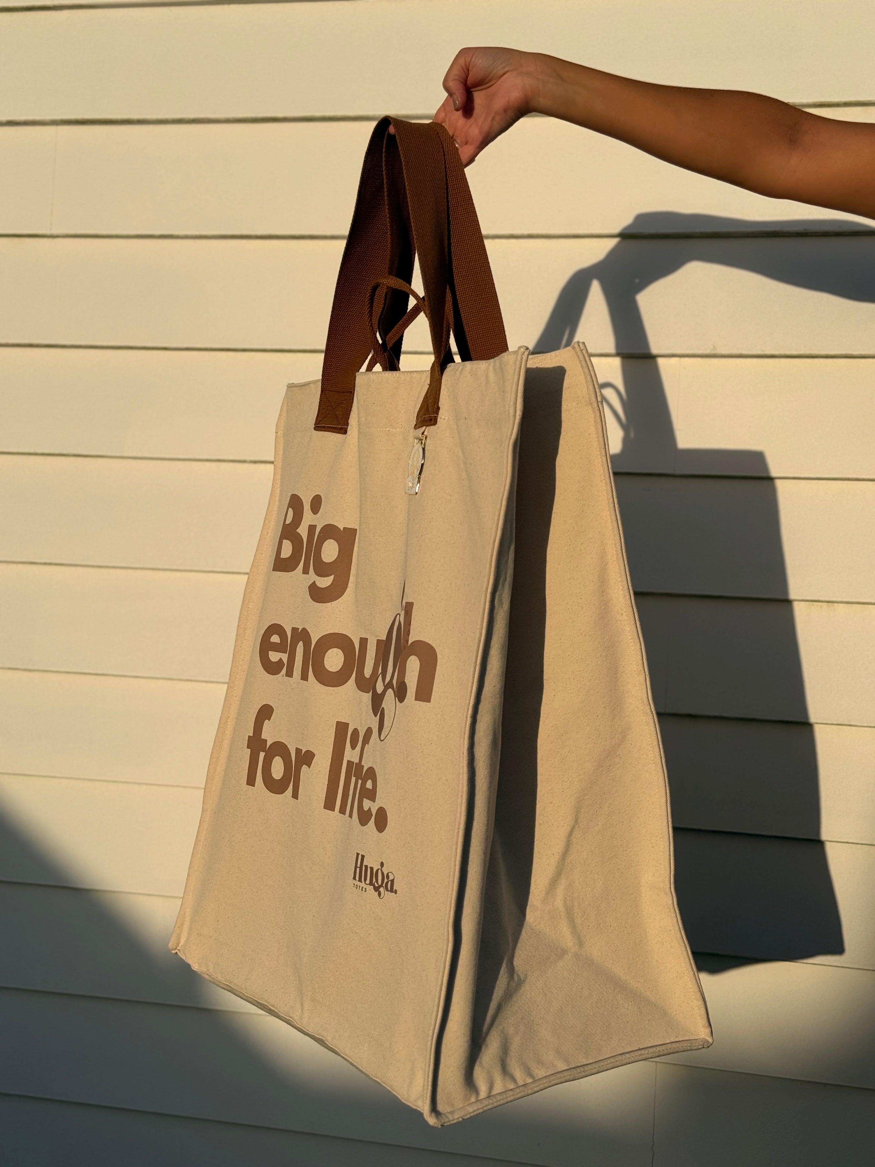 Beige tote bag with brown handles and text 'Big enough for life.' held by a hand against a wooden background.